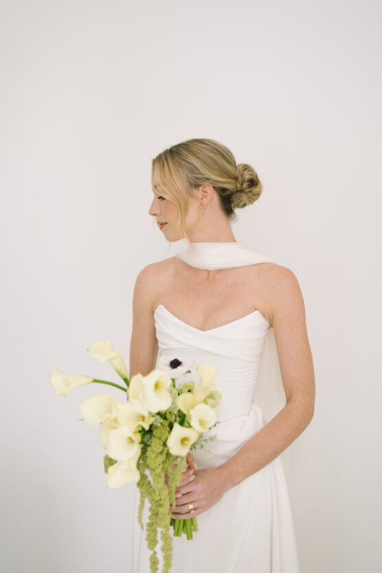 A bride in a white dress with a scarf-like neckline holds a bouquet of white calla lilies. She has her blonde hair in a low bun and is looking to the side against a plain white background. by Justin Salem Meyer
