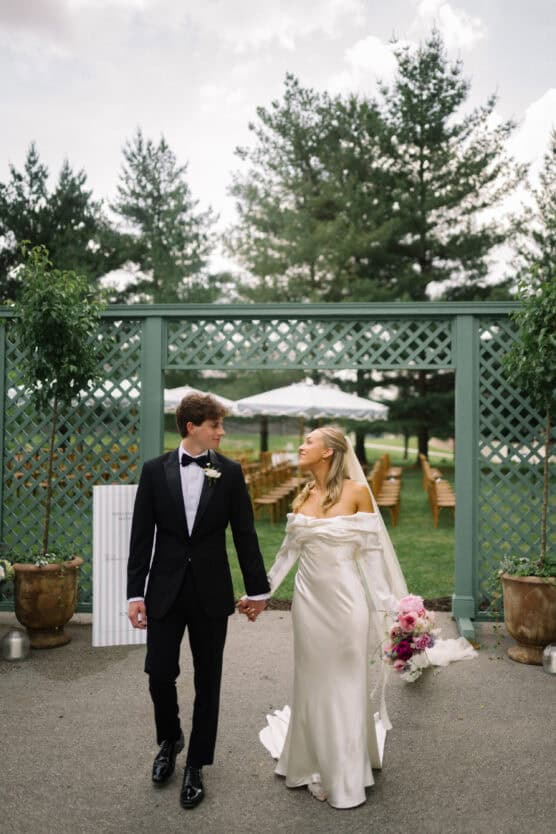 A bride in an off-the-shoulder white gown holds a bouquet and walks hand-in-hand with a groom in a black tuxedo outdoors, with greenery and wedding seating in the background. by Justin Salem Meyer