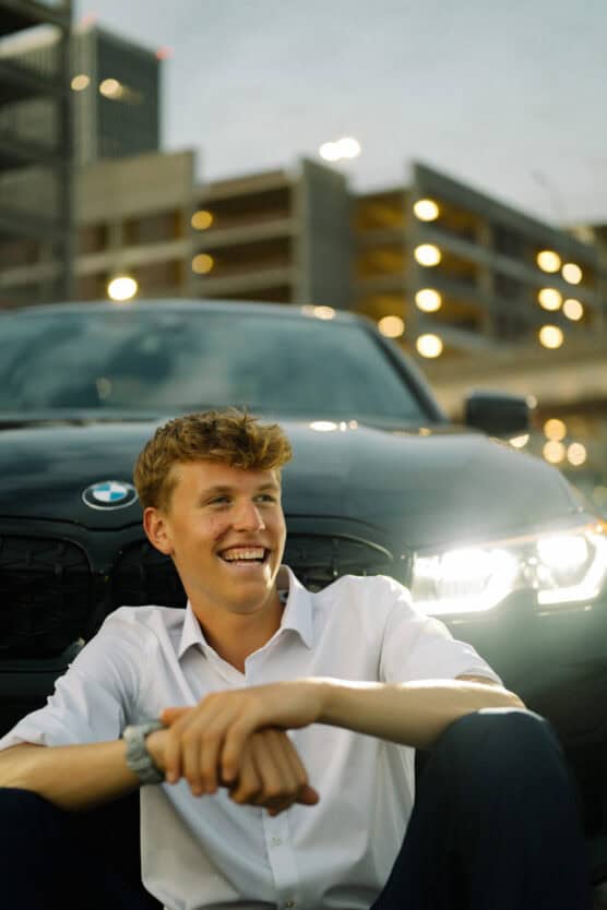 A young man in a white shirt sits smiling in front of a black BMW with its headlights on, with a blurred parking garage and city lights in the background. by Justin Salem Meyer