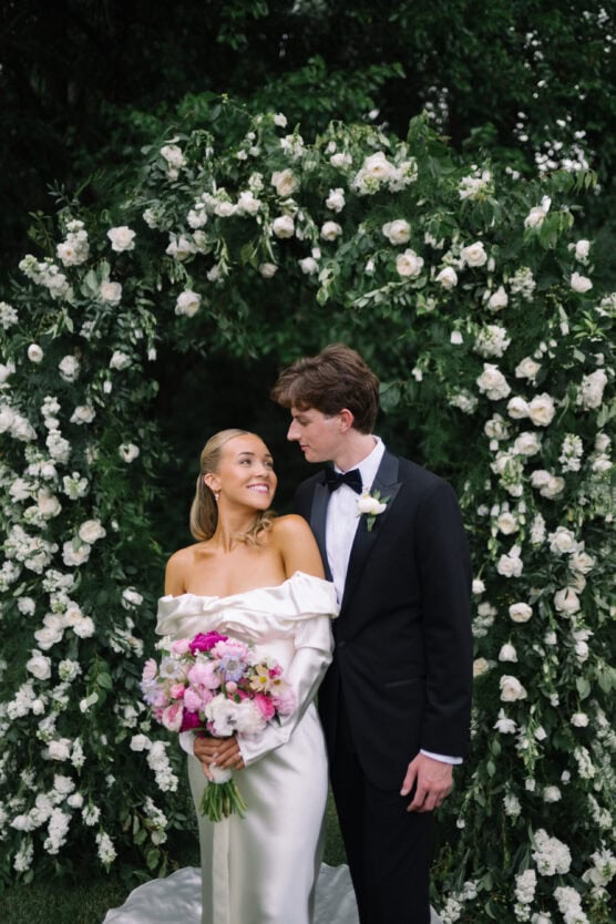 A bride in an off-the-shoulder white gown holding a bouquet of flowers stands smiling beside a groom in a black tuxedo, with a floral arch of white roses and greenery in the background. by Justin Salem Meyer