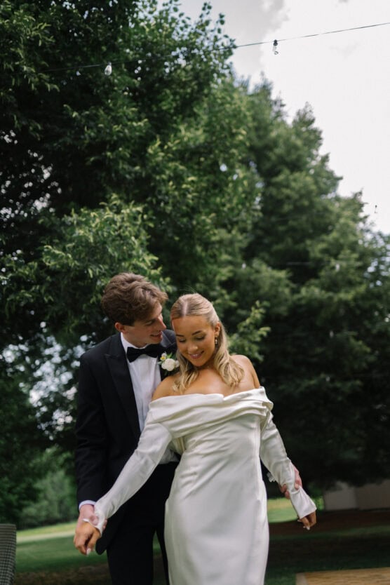 A couple dressed in formal wedding attire smiles and holds hands outdoors, surrounded by lush green trees. The bride wears an off-shoulder white gown, and the groom is in a black tuxedo. by Justin Salem Meyer