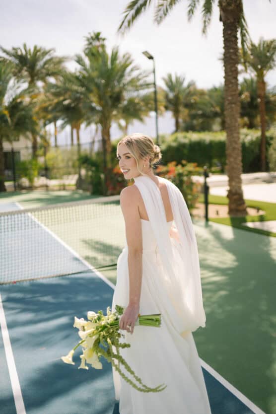 A bride in a white dress and veil holds a bouquet of flowers while walking across a tennis court, looking back over her shoulder. Palm trees and greenery are visible in the background. by Justin Salem Meyer