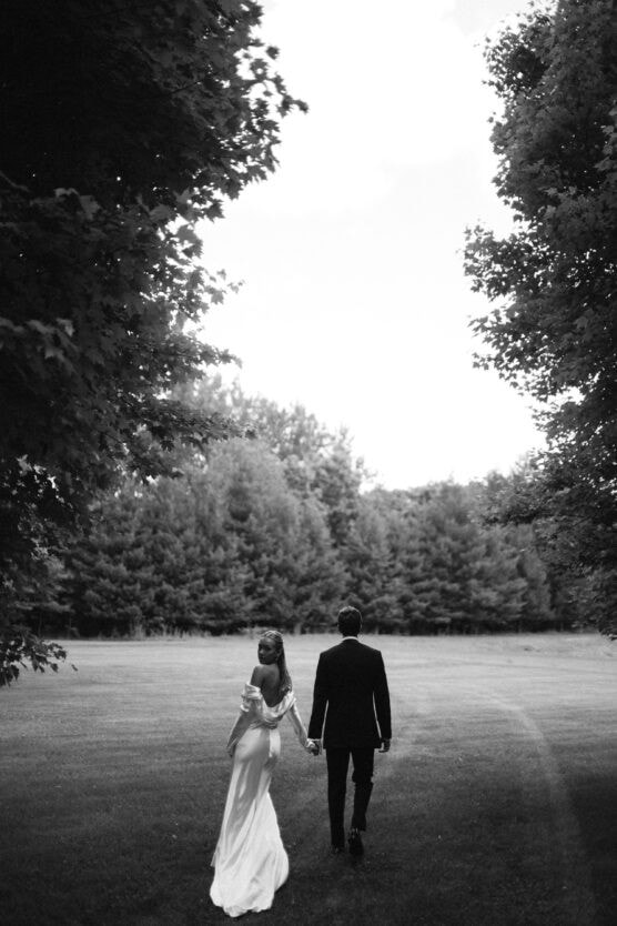 A couple dressed formally, with the woman in a white dress and the man in a dark suit, walk hand in hand across a grassy field surrounded by trees. The scene is serene and captured in black and white. by Justin Salem Meyer