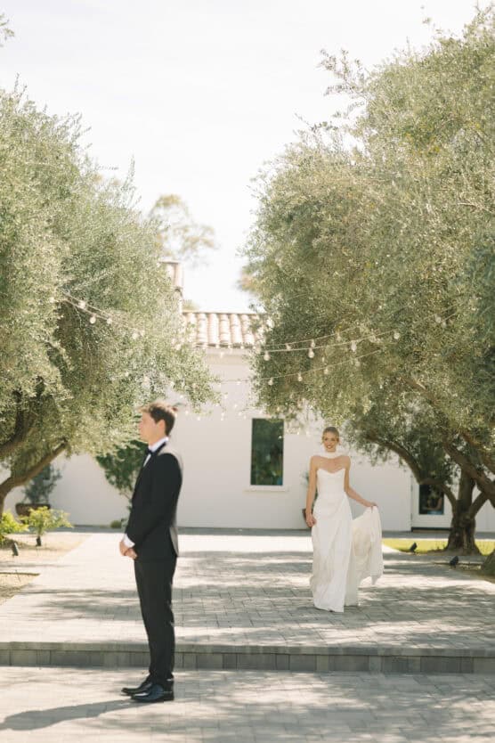 A bride in a white dress walks toward a groom in a black suit, who stands with his back to her, under trees with string lights in a sunlit outdoor setting. by Justin Salem Meyer