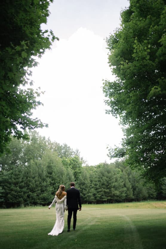 A couple, dressed formally, walk hand-in-hand across a green field bordered by tall trees under a cloudy sky. The scene is peaceful and romantic. by Justin Salem Meyer