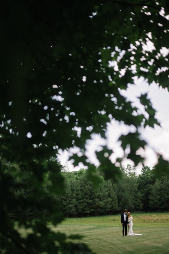 A bride and groom stand together on a grassy field, surrounded by trees, framed by out-of-focus green leaves in the foreground. by Justin Salem Meyer