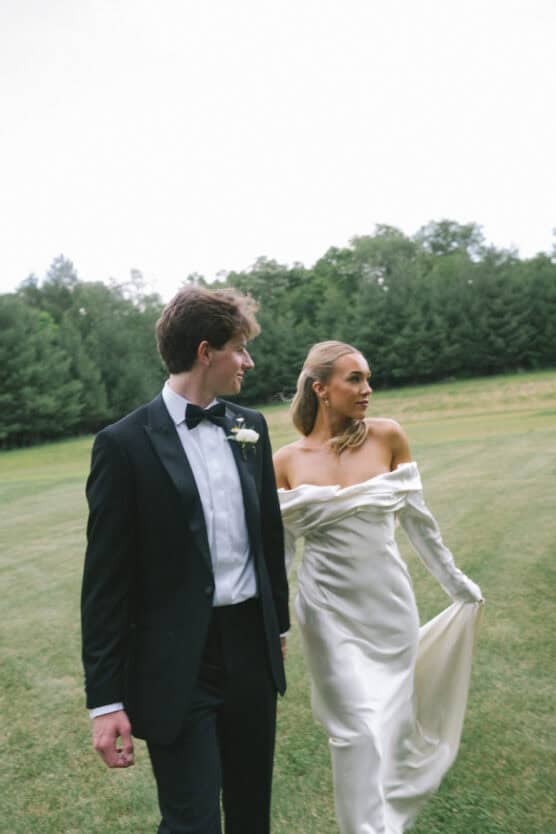 A bride in an off-the-shoulder white gown and a groom in a black tuxedo walk hand in hand across a grassy field, with trees in the background. by Justin Salem Meyer