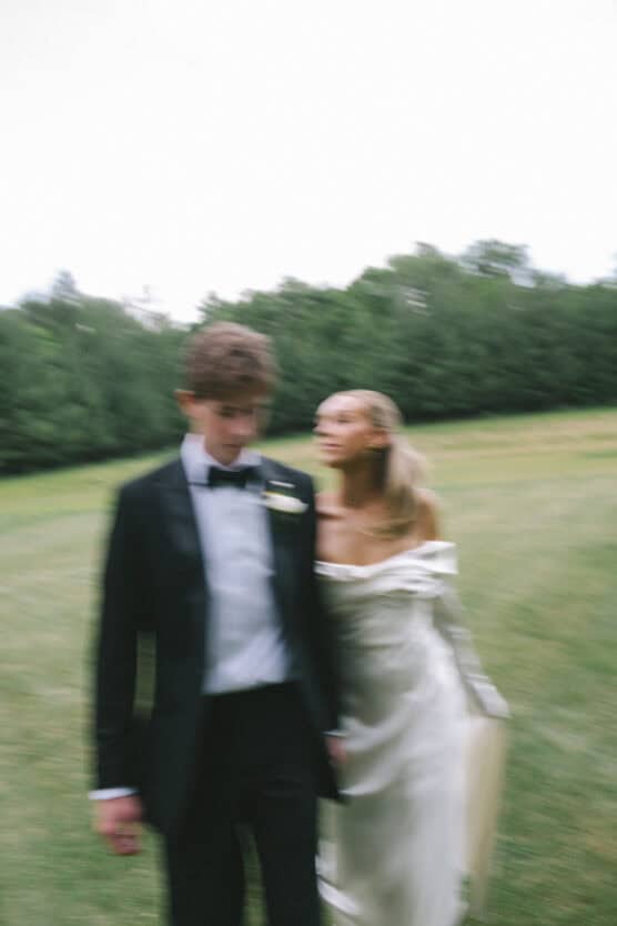 A blurred image of a couple outdoors, with the man in a black tuxedo and the woman in an off-shoulder white dress, standing on grass with trees in the background. by Justin Salem Meyer