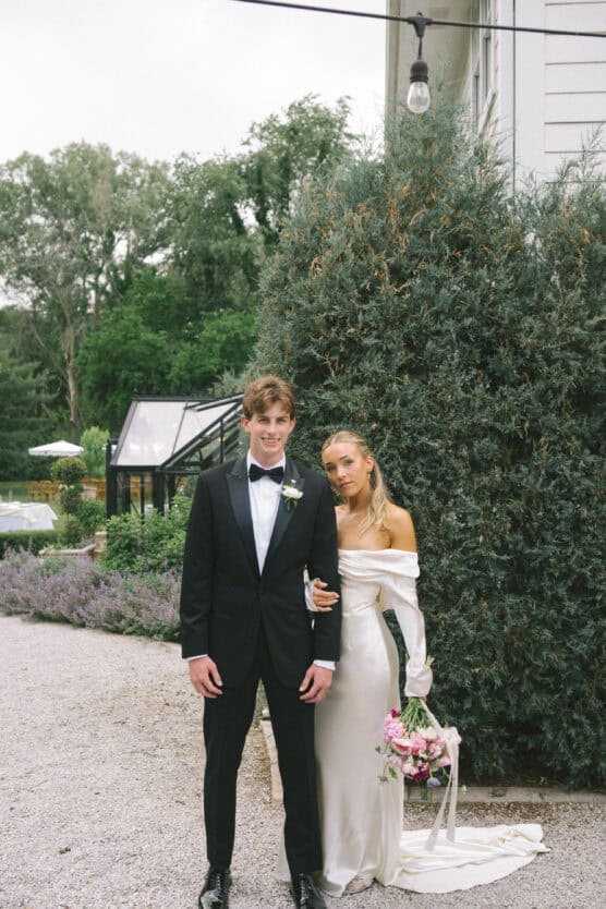 A young couple poses outdoors, dressed formally; the man wears a black tuxedo with a bow tie, and the woman wears an off-shoulder white gown, holding a bouquet of pink flowers. They stand on a gravel path near greenery. by Justin Salem Meyer