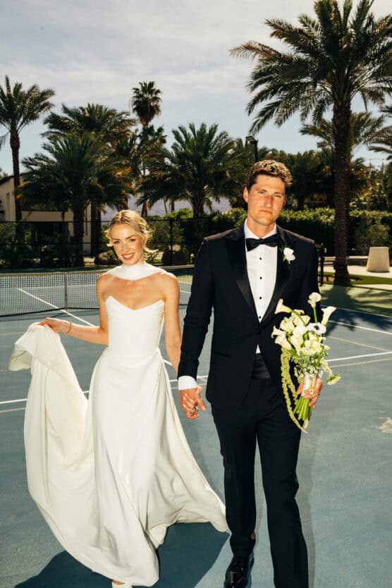 A bride in a white dress and groom in a black tuxedo hold hands and flowers, smiling, walking outdoors on a sunny day with palm trees and a tennis court in the background. by Justin Salem Meyer