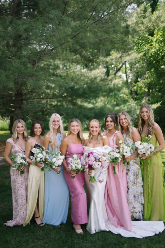 Seven women in colorful dresses stand outdoors on grass, holding bouquets of flowers. They are smiling and posing together, with green trees and sunlight in the background. by Justin Salem Meyer