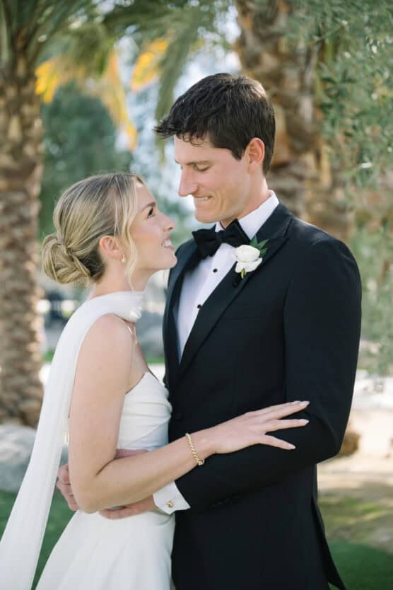 A bride and groom stand close together outdoors, gazing into each others eyes and smiling. The bride wears a white dress and the groom is in a black tuxedo with a white boutonniere; palm trees are in the background. by Justin Salem Meyer