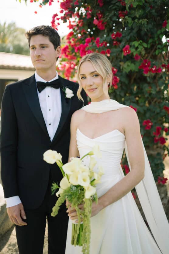A bride in a white gown holding a bouquet of white flowers stands beside a groom in a black tuxedo. They are outdoors, in front of a lush bush with vibrant red blooms. Both have calm, serene expressions. by Justin Salem Meyer