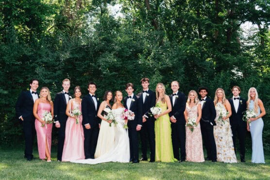 A group of 15 people dressed in formal attire pose outdoors in front of greenery; women wear colorful dresses and hold bouquets, while men wear black tuxedos with bowties. by Justin Salem Meyer