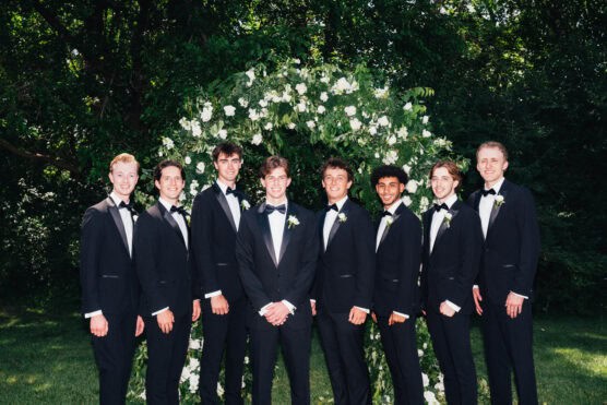 Eight young men in black tuxedos and bow ties stand smiling in front of a green arch decorated with white flowers, outdoors on a sunny day with trees in the background. by Justin Salem Meyer
