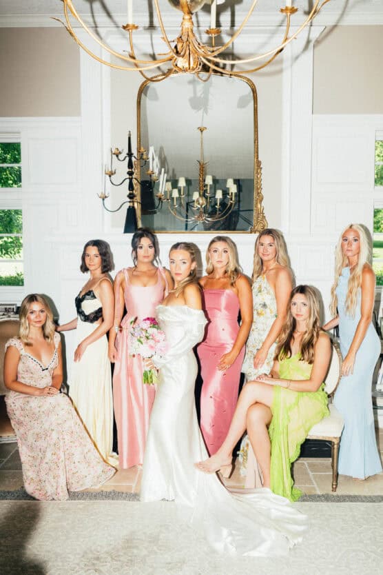 Eight women dressed in elegant, pastel-colored gowns pose together indoors; one in a white dress holds a bouquet, likely the bride, while others stand and sit around her under a chandelier in a bright, stylish room. by Justin Salem Meyer