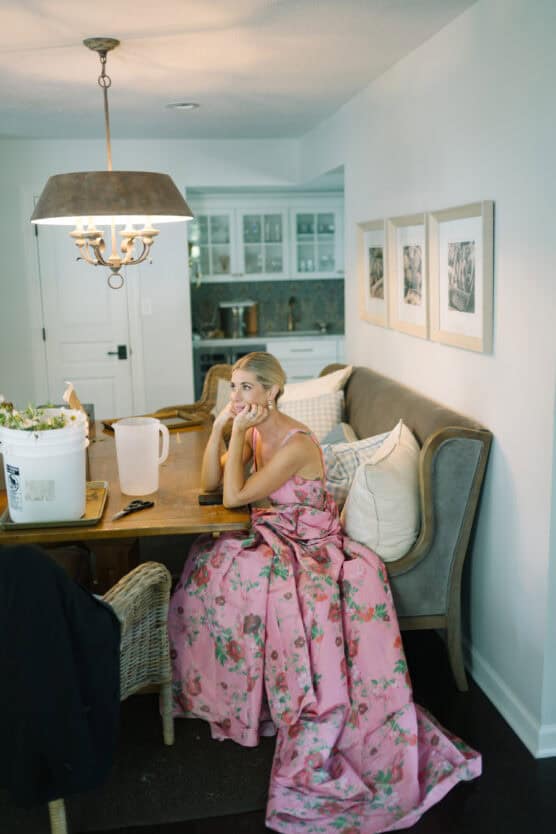 A woman in a pink, floral gown sits thoughtfully at a dining table in a cozy kitchen, resting her chin on her hand. The table has a white pitcher, a large bucket, and a jacket draped over a chair. by Justin Salem Meyer