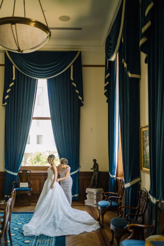 A bride in a white gown stands near a large window with blue curtains, while another woman helps adjust her dress. The elegant room features classic furniture, a chandelier, and artwork on the walls. by Justin Salem Meyer
