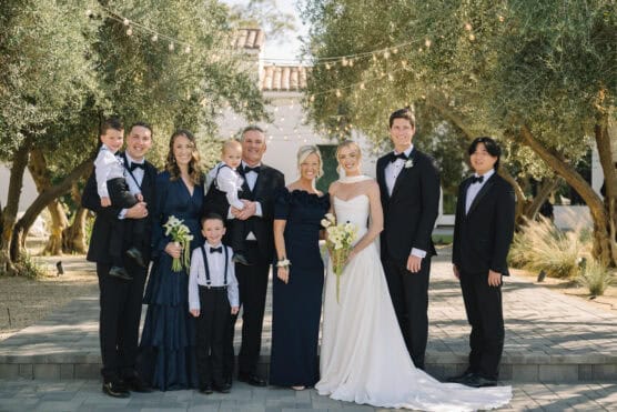 A wedding party poses outdoors on a stone path under trees with string lights. The bride in a white dress stands beside the groom in a tuxedo, surrounded by family in formal navy and black attire, including children. by Justin Salem Meyer