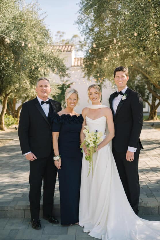 Four people pose outdoors for a formal wedding photo. Two men in black tuxedos and two women in elegant dresses, one in a white bridal gown holding a bouquet, stand together, smiling under string lights and trees. by Justin Salem Meyer