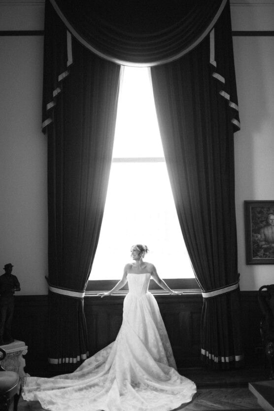 A bride in a flowing gown stands in front of a large, bright window framed by tall, dark curtains. The room has elegant decor and wood paneling, with a portrait and a figure in the background. The image is in black and white. by Justin Salem Meyer