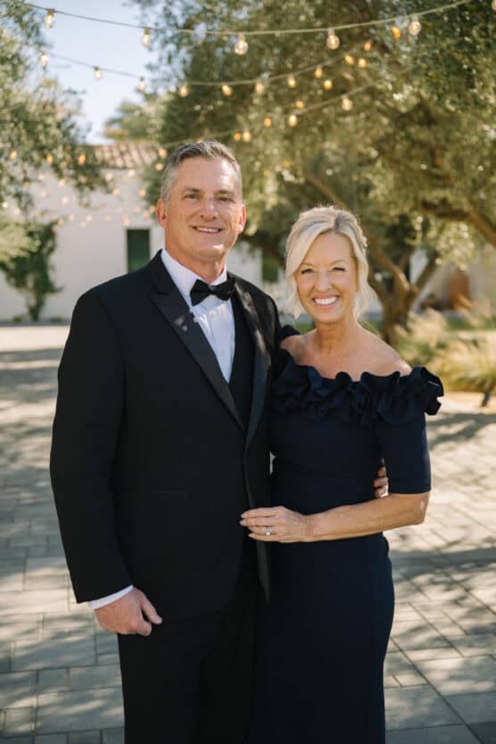 A smiling man in a black tuxedo and a woman in an off-the-shoulder navy dress pose together outdoors under string lights, with trees and a white building in the background. by Justin Salem Meyer