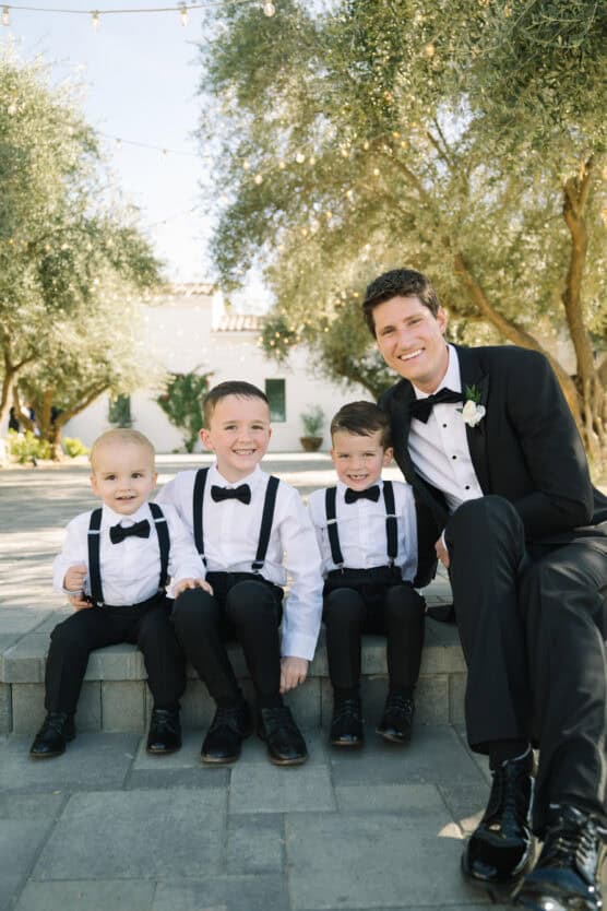 A man in a black tuxedo sits outdoors on stone steps with three young boys, all wearing white shirts, black pants, bow ties, and suspenders. They are smiling and surrounded by trees with string lights above. by Justin Salem Meyer