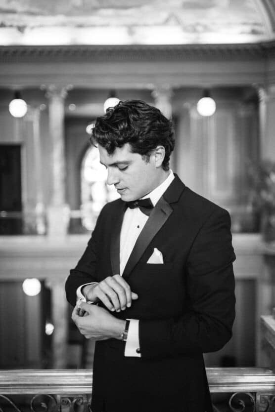 A young man in a tuxedo adjusts his cufflink, standing indoors in an elegant, ornately decorated venue with columns and soft lighting. The image is in black and white. by Justin Salem Meyer