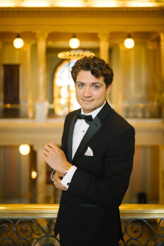 A young man in a black tuxedo with a bow tie stands indoors, smiling slightly. He is in an elegant, ornate building with warm lighting, marble columns, and a decorative railing in the foreground. by Justin Salem Meyer