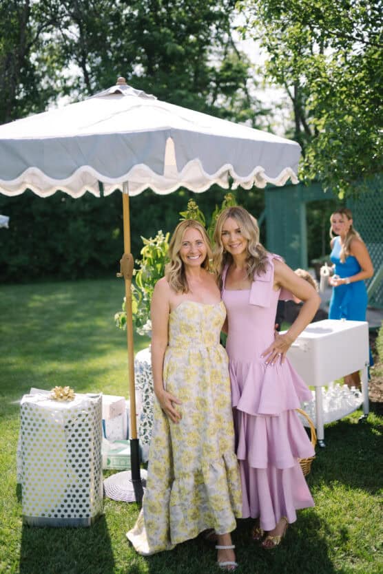 Two women in floral and pink dresses stand smiling together under a white patio umbrella outdoors, surrounded by gift-wrapped presents, with another woman in blue in the background. The setting is a sunny, green garden. by Justin Salem Meyer