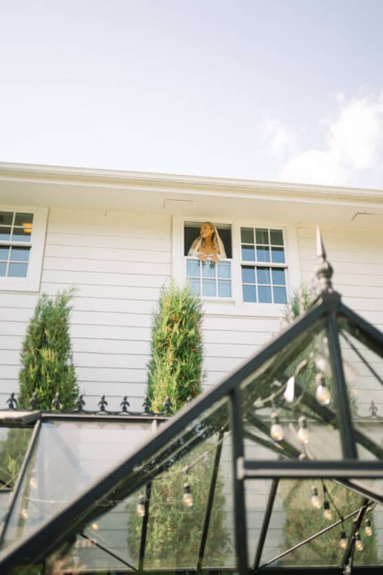 A woman leans out of an open second-story window of a white house, looking up. Tall green bushes stand below, and string lights hang from a glass structure in the foreground. by Justin Salem Meyer
