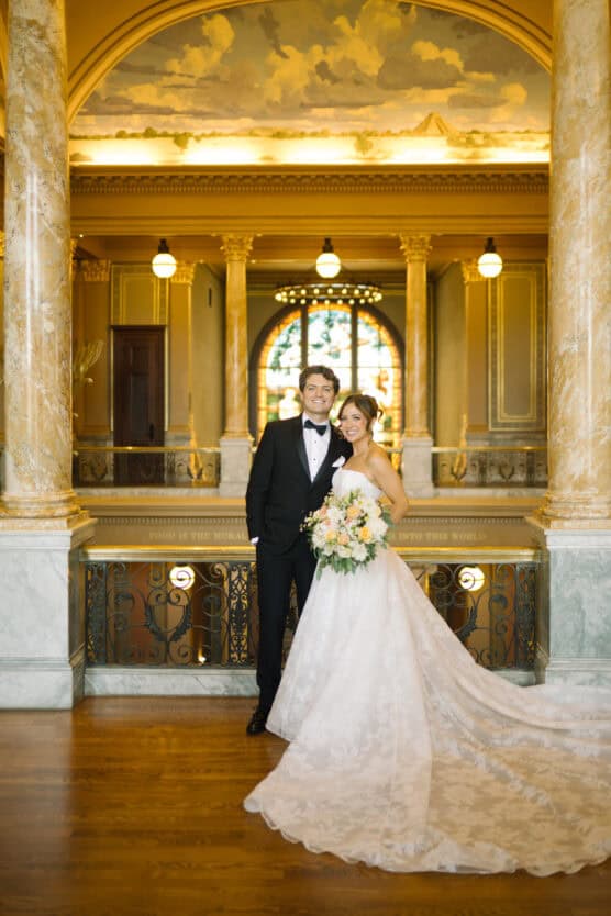 A bride in a white gown holding a bouquet stands beside a groom in a black tuxedo. They smile in an elegant hall with marble columns, ornate railings, and a large arched window in the background. by Justin Salem Meyer