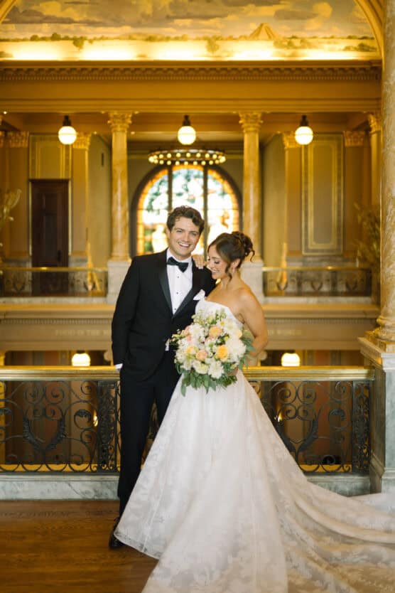 A bride in a white gown holds a bouquet and stands next to a groom in a black tuxedo. They smile at each other in an elegant, ornately decorated hall with columns, warm lighting, and a large arched window in the background. by Justin Salem Meyer