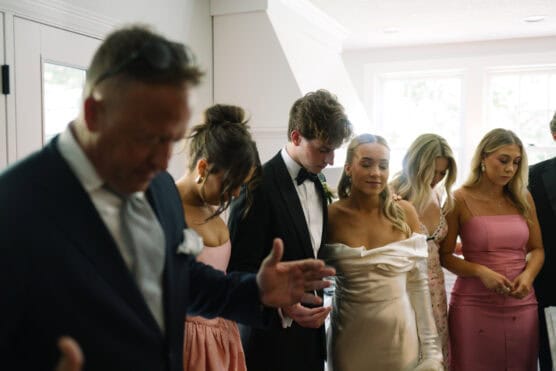 A group of people, including a bride in an off-shoulder gown and a groom in a tuxedo, stand close together in a well-lit room, holding hands and bowing their heads in a moment of prayer or reflection. by Justin Salem Meyer