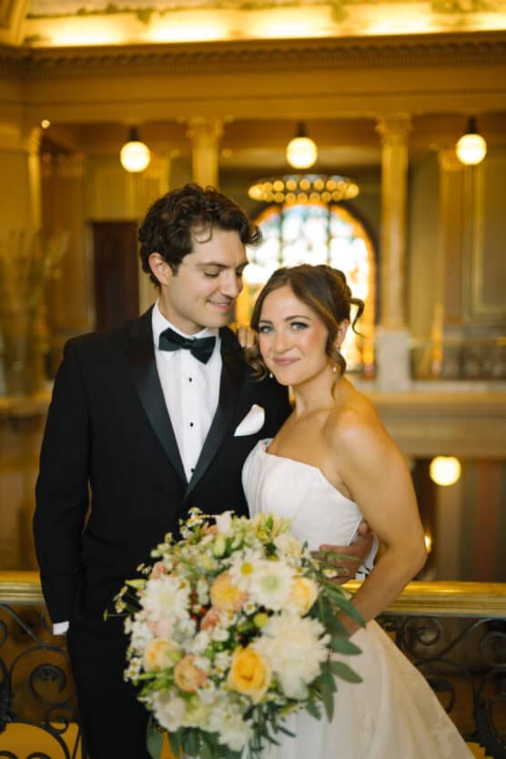 A bride in a strapless white gown holds a bouquet of flowers and stands beside a groom in a black tuxedo, both smiling in an elegant, warmly lit hall with golden decor and arched lights in the background. by Justin Salem Meyer
