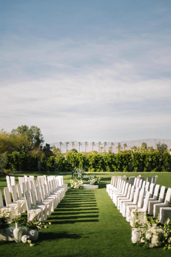 Rows of white chairs set up on a green lawn for an outdoor wedding ceremony, with white floral arrangements lining the aisle and trees and palm trees visible in the background under a blue sky. by Justin Salem Meyer