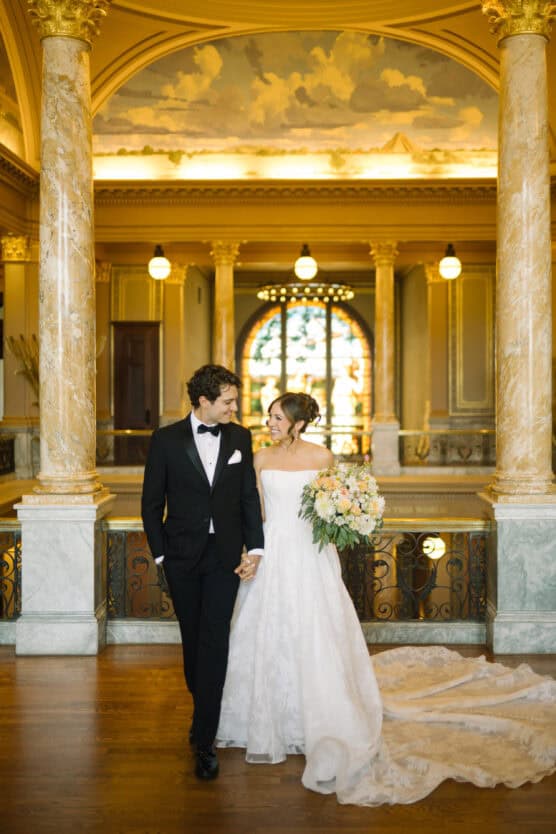 A bride and groom smile at each other while holding hands in an elegant hall with marble columns, ornate railings, and warm lighting. The bride wears a white off-the-shoulder gown and holds a large bouquet. by Justin Salem Meyer