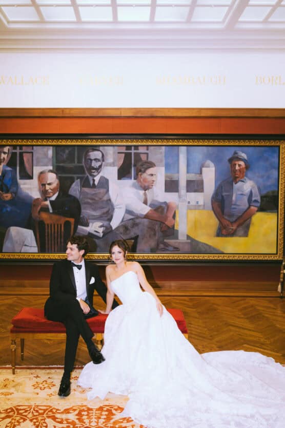 A bride in a white gown and a groom in a black tuxedo sit on a red bench in front of a large mural featuring several painted figures in a formal, elegant room with wood flooring and decorative rug. by Justin Salem Meyer