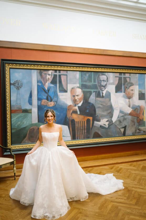 A bride in a white strapless gown stands smiling in front of a large framed painting of four men in a museum or gallery with wood floors and high ceilings. by Justin Salem Meyer