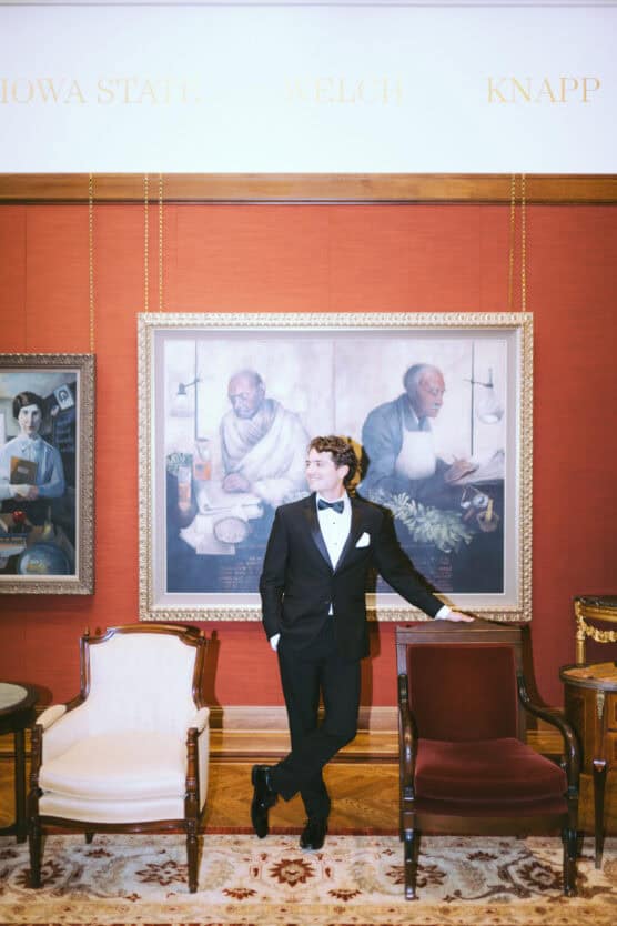 A man in a black tuxedo stands between two chairs, leaning on one, in front of a large framed painting and a red wall. The setting appears formal and elegant with portraits and ornate furniture. by Justin Salem Meyer