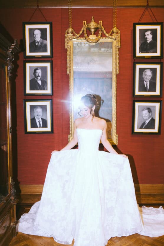 A woman in a strapless white wedding dress stands indoors, holding out her gown. A bright camera flash obscures her face. She is in front of a large ornate mirror and surrounded by framed portraits on a red wall. by Justin Salem Meyer