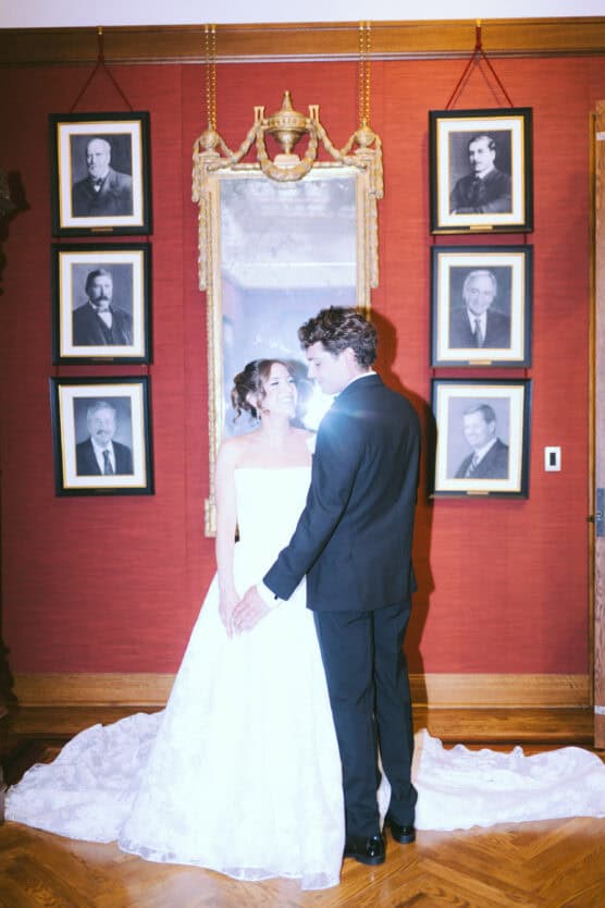 A bride in a white gown and a groom in a black suit stand together, smiling and holding hands, in front of a red wall adorned with framed black-and-white portraits and a large ornate mirror. by Justin Salem Meyer
