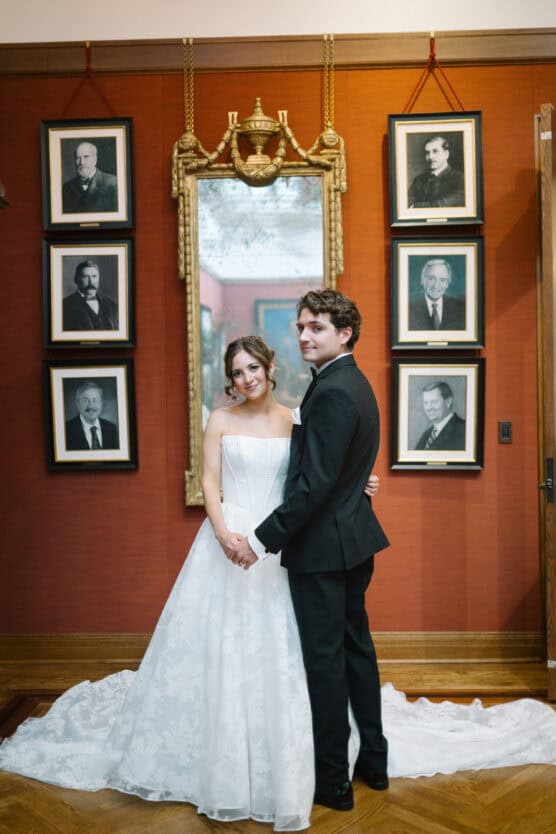 A bride in a white gown and a groom in a black tuxedo stand smiling together in front of a large mirror, surrounded by framed portraits on a reddish-brown wall. by Justin Salem Meyer