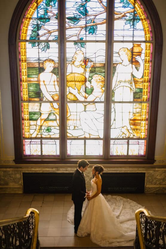 A bride and groom stand holding hands at the base of a staircase, looking at each other, with a large, colorful stained glass window featuring four figures as the backdrop. by Justin Salem Meyer