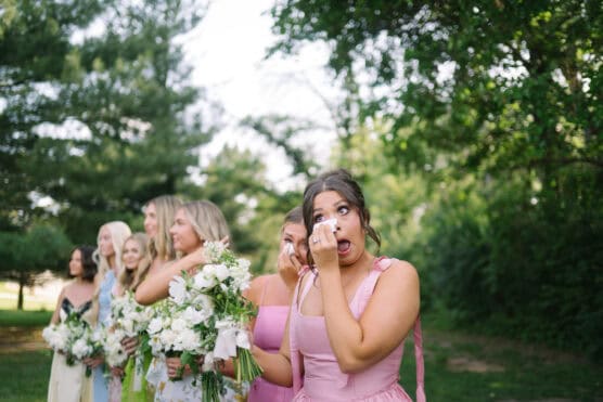 A woman in a pink dress wipes her tears while standing outdoors with other bridesmaids holding bouquets. The group is surrounded by greenery and trees, suggesting a wedding or special event. by Justin Salem Meyer