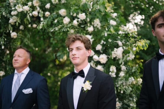 Three men dressed formally in suits and tuxedos stand outdoors in front of a lush green arch covered in white flowers, likely at a wedding or formal event. The central figure wears a black tuxedo with a boutonniere. by Justin Salem Meyer