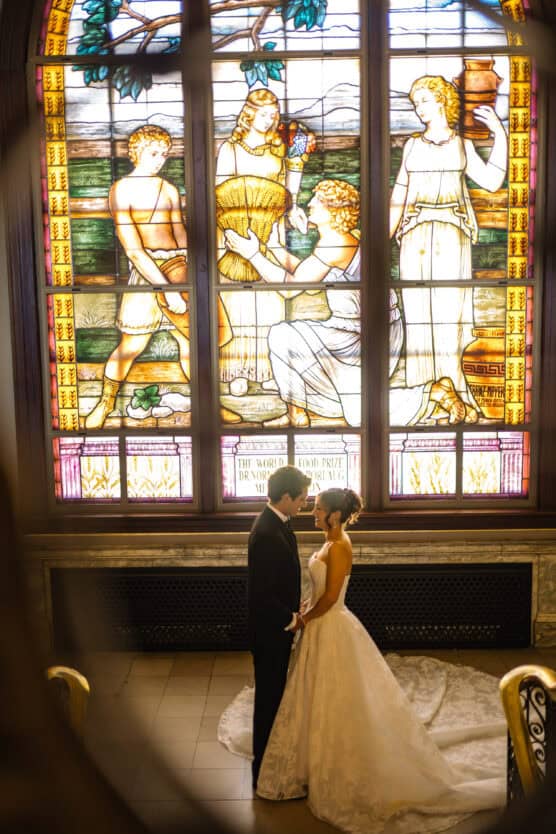 A bride and groom stand together in front of a large, ornate stained glass window featuring figures in classical attire. Warm light filters through, highlighting the couple in wedding attire. by Justin Salem Meyer