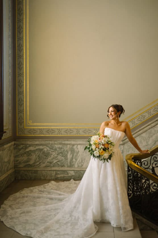 A bride in a strapless white gown with a long train stands on a marble staircase, holding a bouquet of flowers and smiling to the side in a softly lit, elegant setting. by Justin Salem Meyer
