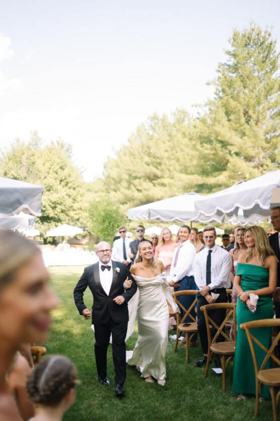 A smiling bride in a white dress walks down an outdoor aisle arm-in-arm with an older man in a tuxedo, surrounded by standing guests and greenery on a sunny day. by Justin Salem Meyer