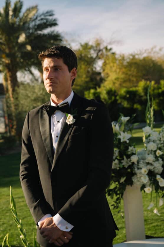 A man in a black tuxedo stands outdoors, hands clasped, with a serious expression. Behind him are green trees, sunlight, and a floral arrangement with white flowers. by Justin Salem Meyer